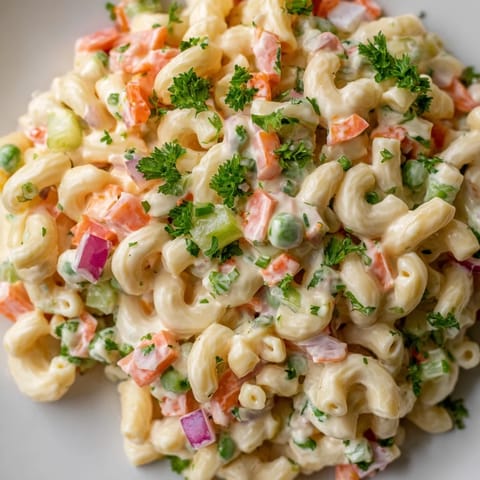 A bowl of creamy macaroni salad filled with diced red bell pepper, peas, and carrots, garnished with fresh chives for a fresh taste.