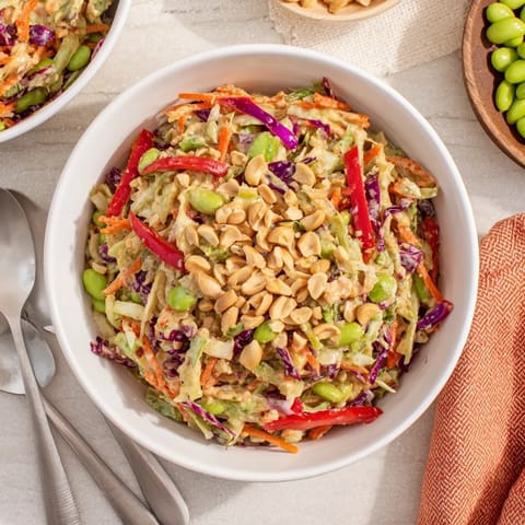 A fresh bowl of Crunchy Thai Peanut Salad, featuring crisp shredded cabbage, carrots, and edamame tossed in a rich peanut dressing, garnished with sesame seeds and lime wedges.