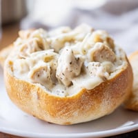 A golden crispy bread bowl holds creamy chicken Alfredo, topped with melted Parmesan cheese for a comforting Italian-American dinner.